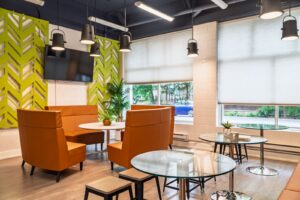 Modern resident lounge with orange booth seating, glass tables, hanging lights, and large windows overlooking greenery.