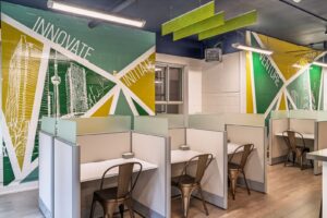 Contemporary workspace of individual study booths with light surfaces, frosted dividers, and bronze café chairs; overhead acoustic panels and industrial lighting; a large green-and-gold mural with city illustrations and motivational words.
